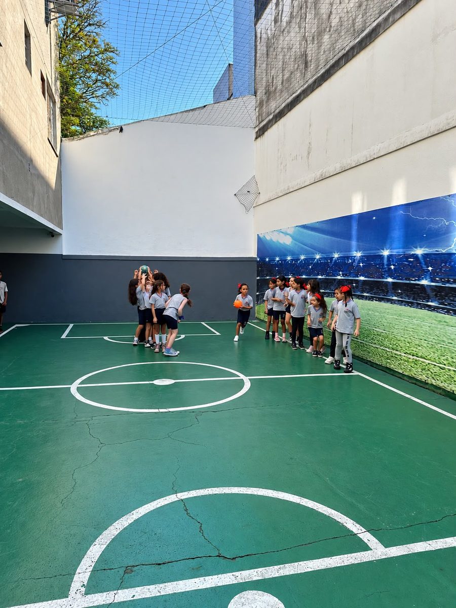 Alunos na quadra poliesportiva coberta com tela de proteção e painel de estádio