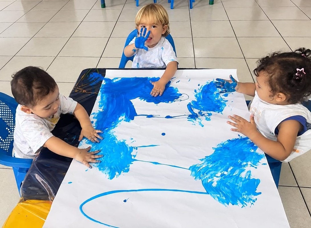 Crianças da Educação Infantil pintando com as mãos em tinta azul sobre um papel grande